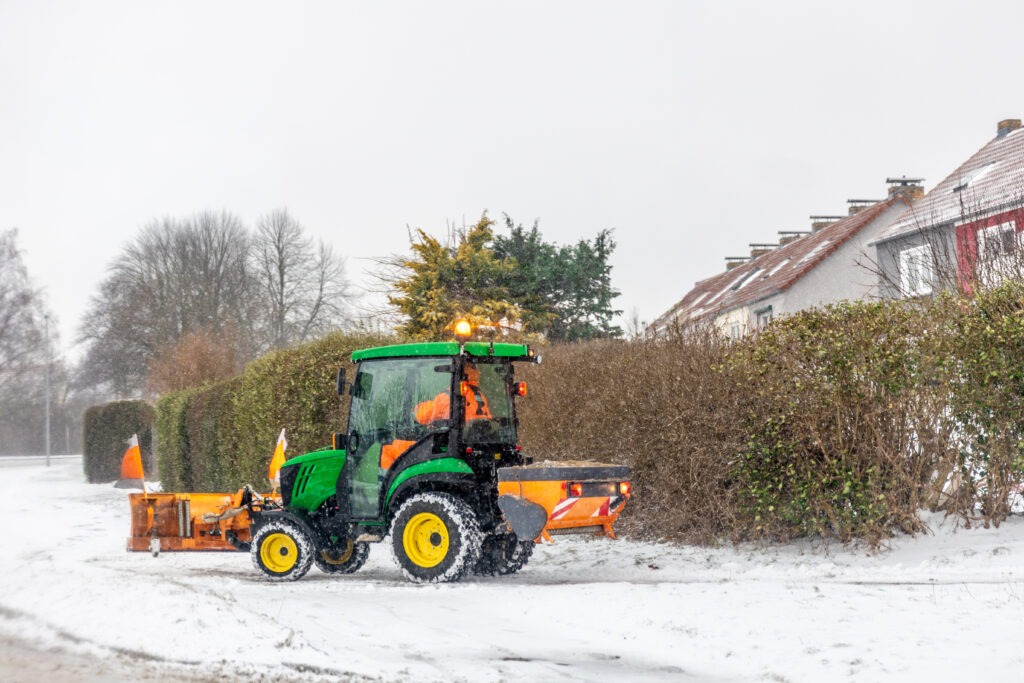 Winterbetreuung: Ein grüner Traktor mit Schneepflug räumt bei leichtem Schneefall eine verschneite Wohnstraße. Die Szene vermittelt die Bedeutung von kommunaler Instandhaltung, Infrastrukturpflege und Wintervorsorge.
